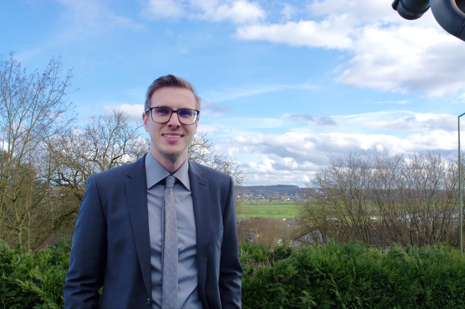 Ein Mann mit Brille und Anzug steht in der Natur. Es ist leicht bewölkt, im Hintergrund sieht man Felder und vereinzelte Bäume.