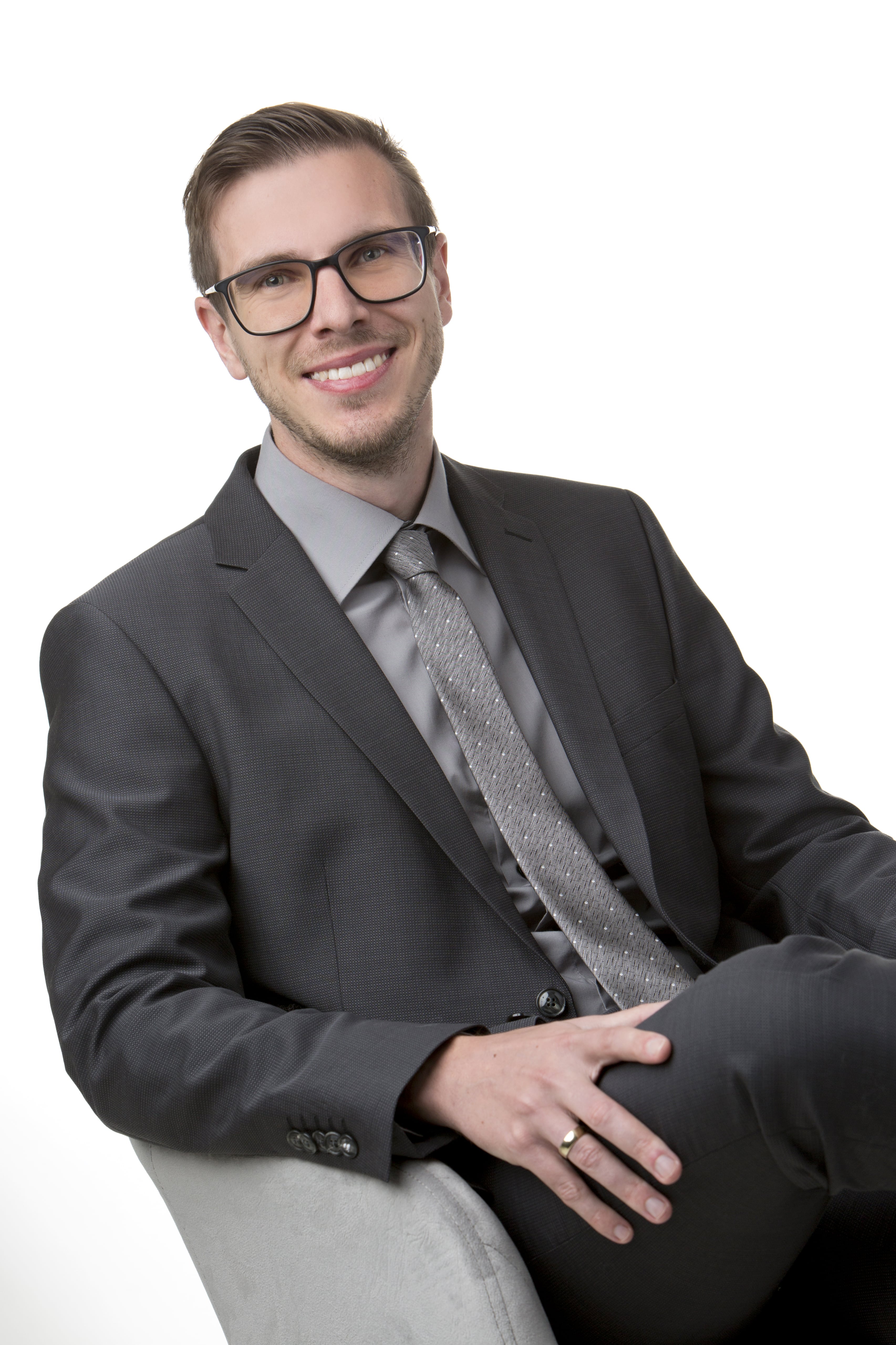 Porträt eines freundlichen Mannes mit Brille, die einen grauen Anzug trägt und in einem grauen Sessel sitzt. Die Beine sind überschlagen.