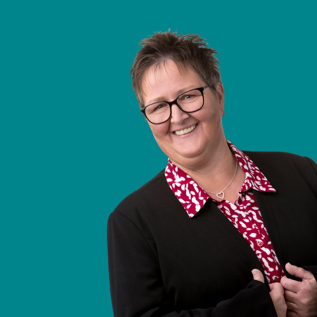 Porträt einer lächelnden Frau mit Kurzhaarschnitt und Brille, die ein rot/weißes Hemd und einen schwarzen Blazer trägt.