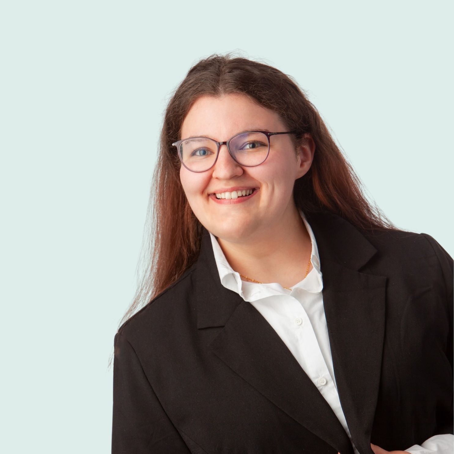 Porträt einer brünetten Frau, die ein weißes Hemd und einen schwarzen Blazer trägt. Der Hintergrund ist in einem hellen blau.