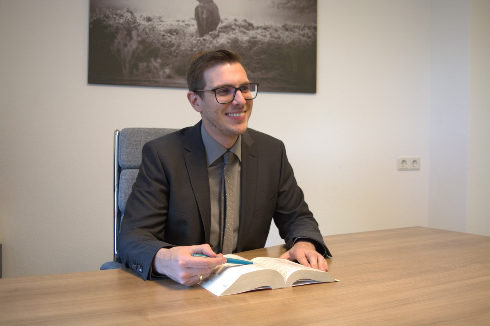 Ein Mann mit Brille und Anzug sitzt an einem Holzschreibtisch und blättert in einem Buch.