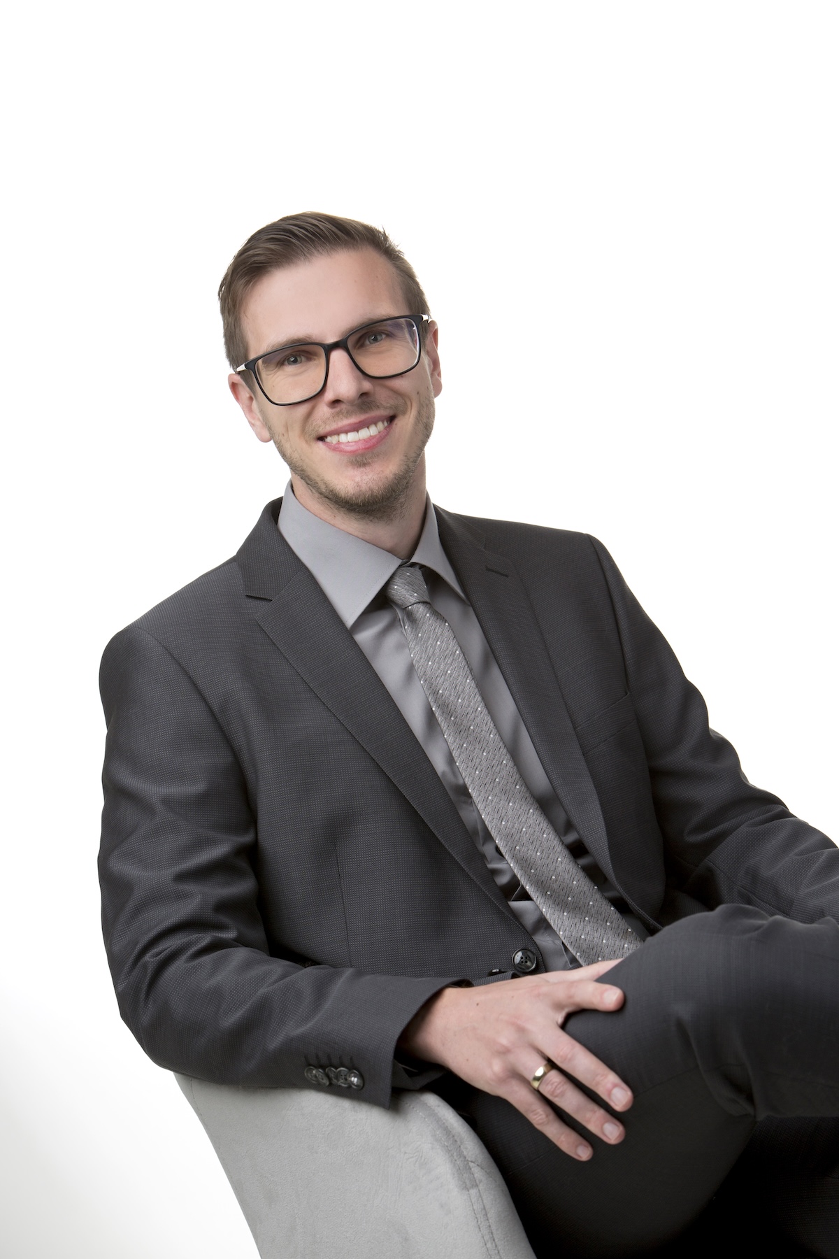 Porträt eines freundlichen Mannes mit Brille, die einen grauen Anzug trägt und in einem grauen Sessel sitzt. Die Beine sind überschlagen.