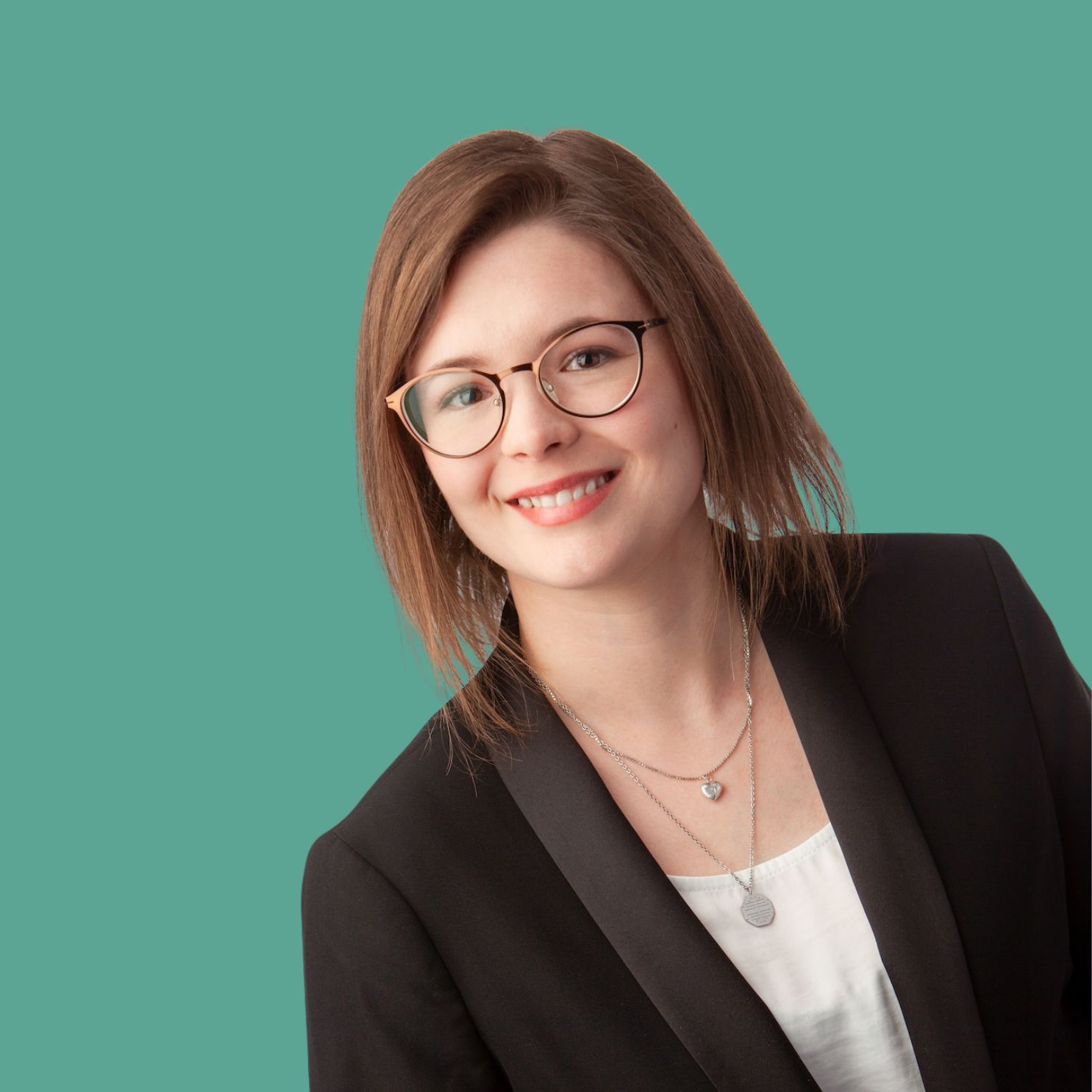 Porträt einer freundlichen, brünetten Frau mit Brille, die ein weißes Shirt und einen schwarzen Blazer trägt. Der Hintergrund ist grün/türkis.