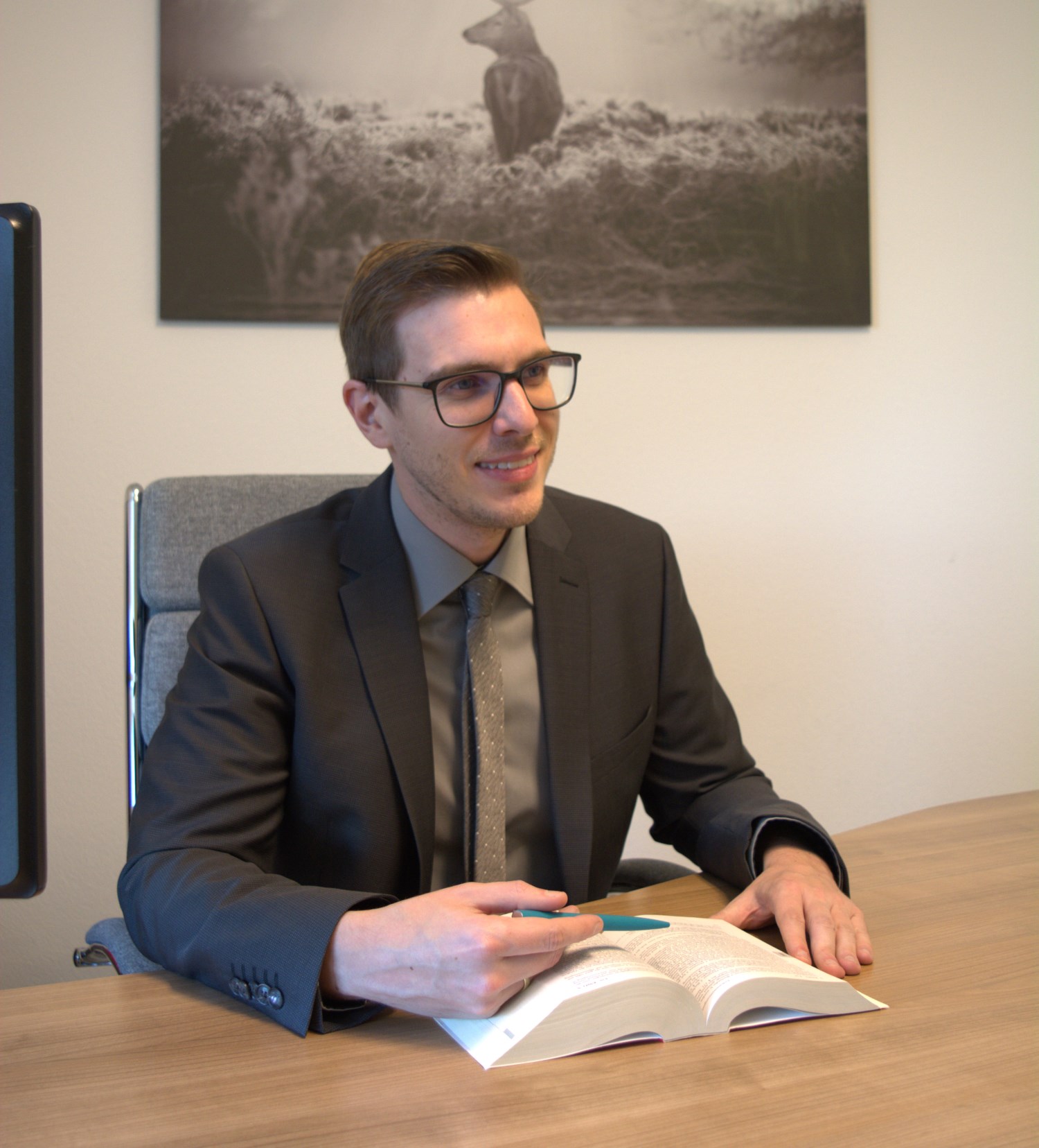 Ein Mann mit Brille und Anzug sitzt an einem Holzschreibtisch und blättert in einem Buch.