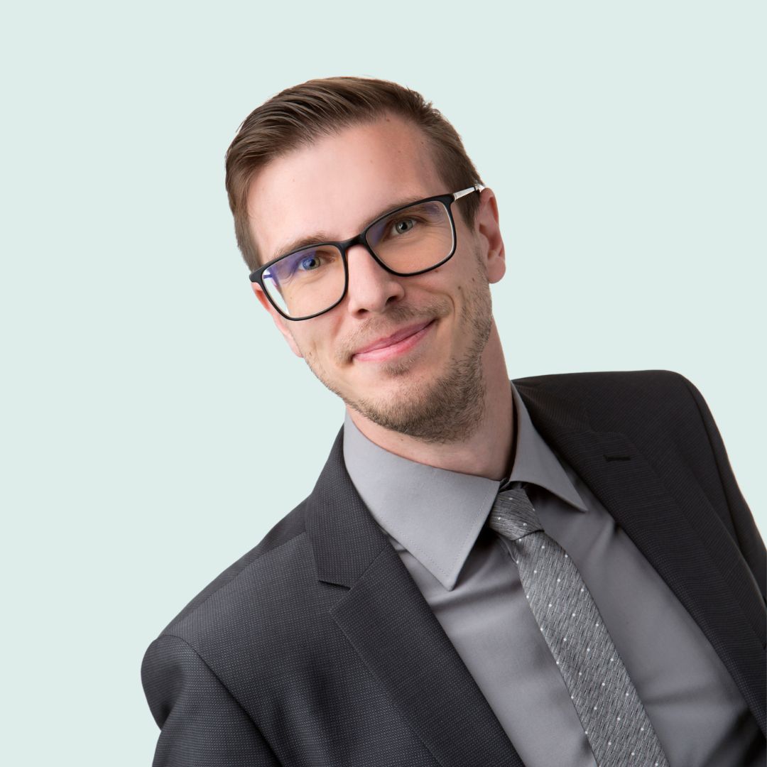 Porträt eines freundlichen Mannes mit Brille, die einen grauen Anzug trägt.